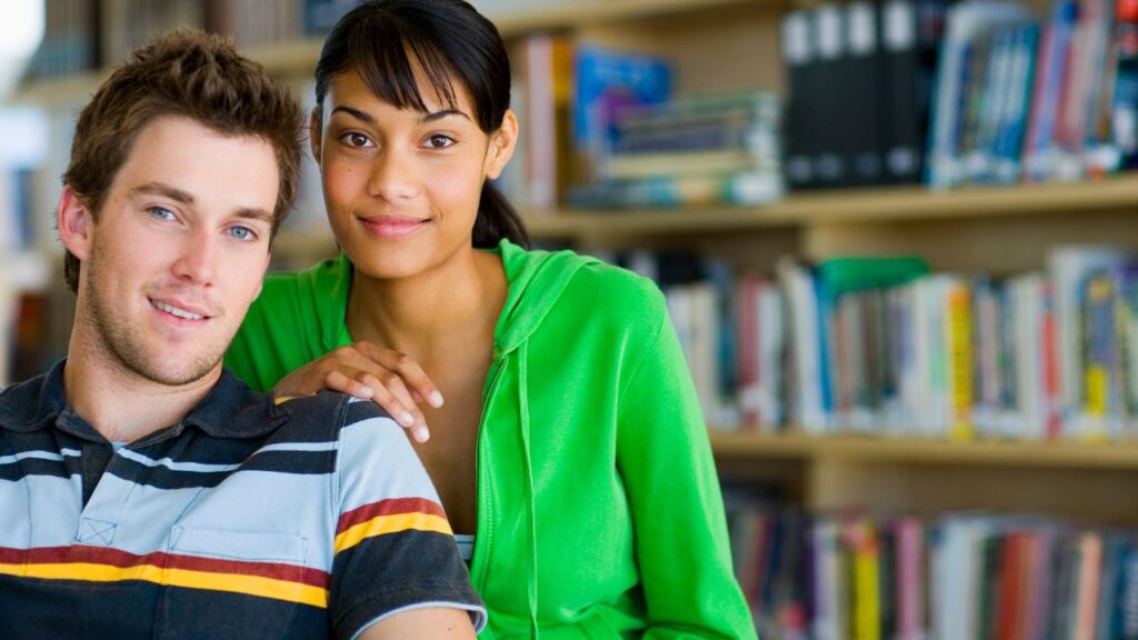 Couple mixte dans une bibliothèque, l'homme regardant la caméra et la femme souriant, évoquant l'importance de la communication pour un homme Verseau.