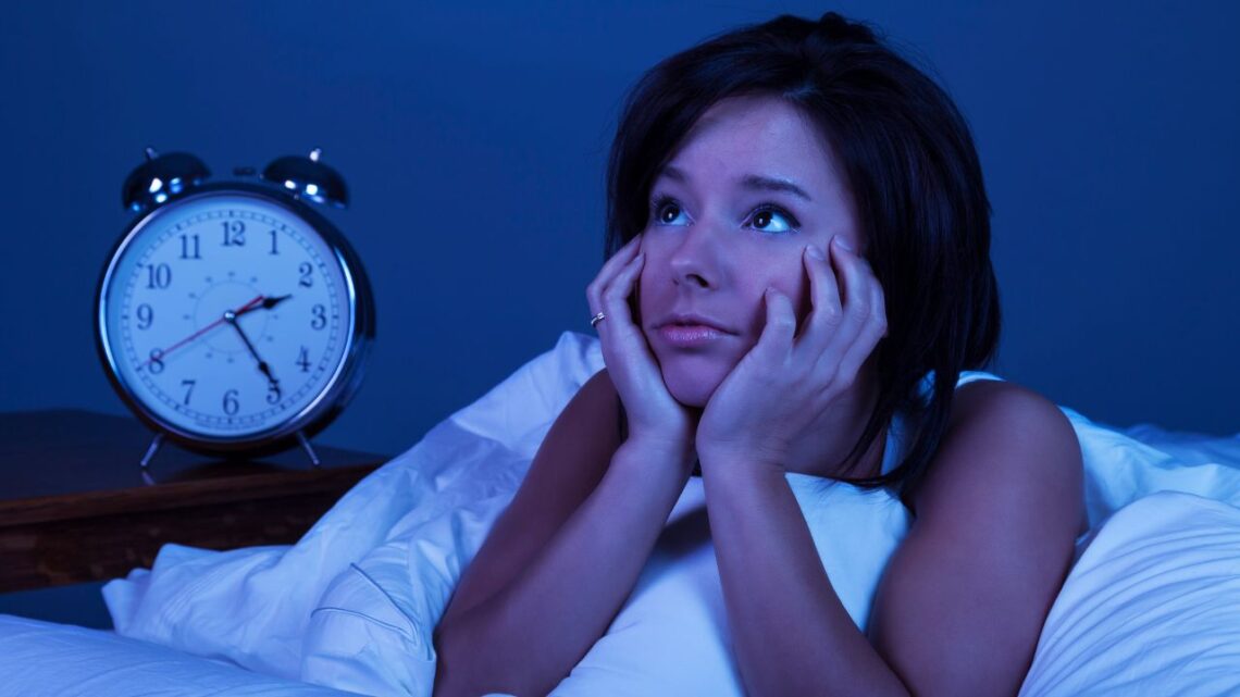 Femme éveillée et inquiète dans son lit à côté d'un réveil indiquant 2 heures.